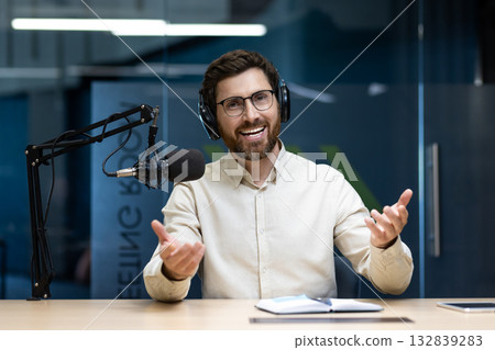 Portrait of a smiling young man in headphones and glasses sitting at a table in front of a microphone and talking to the camera, gesturing with his hands. Portrait of a smiling young man in headphones and glasses sitting at a table in front of a microphone and talking to the camera, gesturing with his hands. 132839283