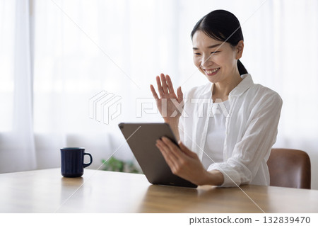 一位面帶微笑的女士在客廳桌子上對著平板電腦揮手。 132839470