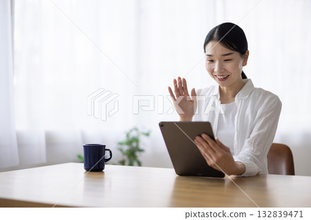 一位面帶微笑的女士在客廳桌子上對著平板電腦揮手。 132839471