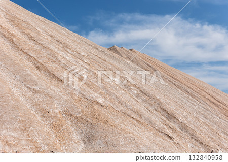 Potassium ore dump. Massive mound potash waste rises deep blue sky, its surface marked by mineral lines erosion patterns. Arid landscape sparse vegetation industrial origin and stark natural contrast 132840958