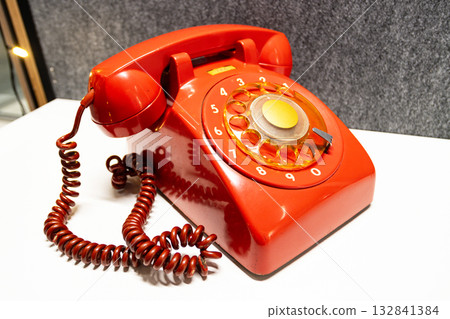 Vintage rotary telephone red retro style. A bright red antique telephone with a gold-tone center dial rests on a white surface, evoking 20th-century communication nostalgia 132841384