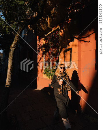Fashionable blonde strolling bistro sidewalk during golden hour, drinking takeaway coffee, wearing long dark coat and leopard print scarf, radiating sophisticated urban style. Stylish blonde woman 132841390