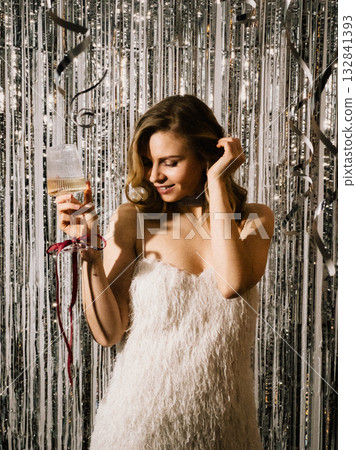 Young woman wearing white dress holding champagne flute and looking at it while touching her hair during New Years Eve party with silver and white metallic streamers hanging on wall. New Years Eve 132841393