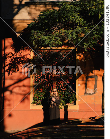 Blonde woman in sunglasses and coat, holding coffee cup, stands near old building with decorative window, bathed in warm glow of afternoon sun. Portrait of blonde woman wearing sunglasses and coat Blonde woman in sunglasses and coat, holding coffee cup, stands near old building with decorative window, bathed in warm glow of afternoon sun. Portrait of blonde woman wearing sunglasses and coat 132841396