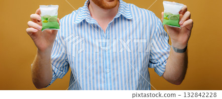 Man in striped shirt holding two green packaged snacks, close-up shot on yellow background Man in striped shirt holding two green packaged snacks, close-up shot on yellow background 132842228