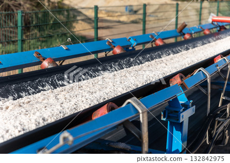 Long blue conveyor belt transports white potash ore material toward massive potassium mining dump sunlight. Industrial machinery stretches distance mineral extraction continuous production process Long blue conveyor belt transports white potash ore material toward massive potassium mining dump sunlight. Industrial machinery stretches distance mineral extraction continuous production process 132842975