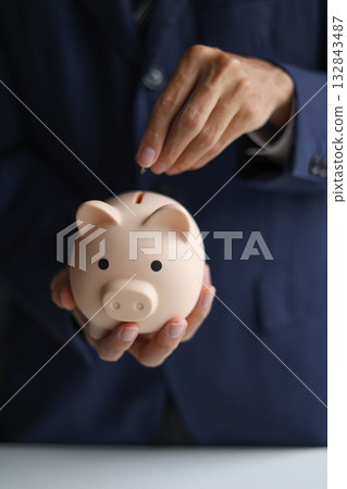A person in a suit putting a coin into a piggy bank. Saving money and financial planning, financial growth, investment, and future planning concept. A person in a suit putting a coin into a piggy bank. Saving money and financial planning, financial growth, investment, and future planning concept. 132843487