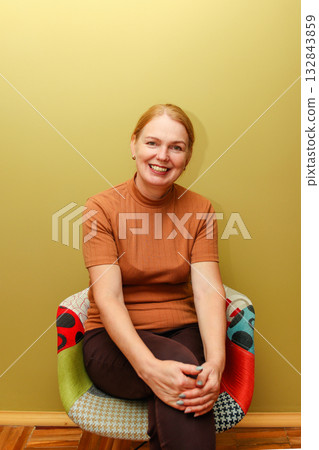 Smiling middle-aged woman with red hair in brown top sits on colorful patchwork chair against light green wall. portrait of a middle-aged woman 45 years old, with a warm smiling expression 132843859