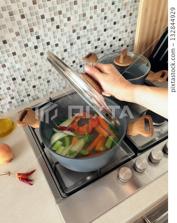 Anonymous person prepares vegetable broth at home in kitchen. Pot with lid on gas stove. Healthy eating. Anonymous person prepares vegetable broth at home in kitchen. Pot with lid on gas stove. Healthy eating. 132844929