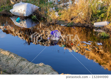 A waterway with dark, polluted water reflects garbage and waste along its banks, highlighting significant environmental issues 132844990