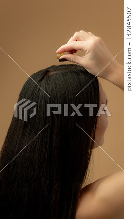Dark haired woman applying vitamin capsule oil to scalp for nourishment and strengthening Dark haired woman applying vitamin capsule oil to scalp for nourishment and strengthening 132845507