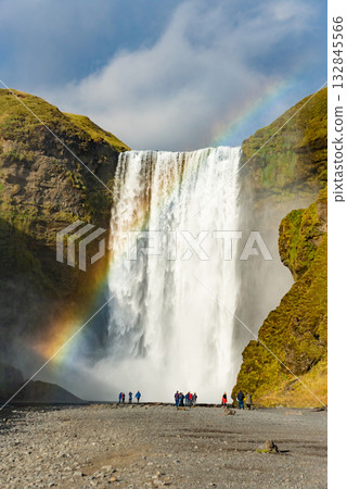 Skogafoss waterfall in Iceland cascades down a steep cliff, creating a powerful mist that forms a vibrant rainbow. The surrounding lush green landscape enhances its majestic beauty Skogafoss waterfall in Iceland cascades down a steep cliff, creating a powerful mist that forms a vibrant rainbow. The surrounding lush green landscape enhances its majestic beauty 132845566
