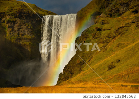 Skogafoss waterfall in Iceland cascades down a steep cliff, creating a powerful mist that forms a vibrant rainbow. The surrounding lush green landscape enhances its majestic beauty Skogafoss waterfall in Iceland cascades down a steep cliff, creating a powerful mist that forms a vibrant rainbow. The surrounding lush green landscape enhances its majestic beauty 132845568
