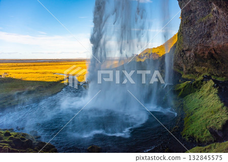 Water cascades from Seljalandsfoss waterfall, creating a misty curtain. The scene captures the lush landscape and stunning blue sky, offering a unique perspective from behind the falls. Water cascades from Seljalandsfoss waterfall, creating a misty curtain. The scene captures the lush landscape and stunning blue sky, offering a unique perspective from behind the falls. 132845575