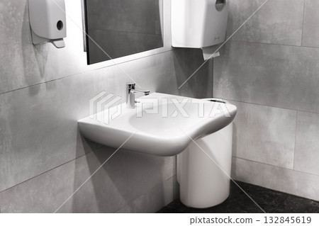 A wide, modern wall-mounted public restroom sink with a short faucet, mirror, soap, and paper dispensers, set against gray tiles. A wide, modern wall-mounted public restroom sink with a short faucet, mirror, soap, and paper dispensers, set against gray tiles. 132845619