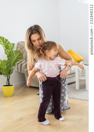Mother helping her daughter get dressed at home with patience and care. Parenting, tenderness, and the everyday love within calm family routine. 132848312