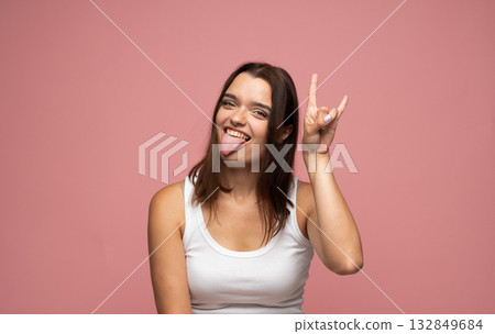 Playful young woman sticking out tongue making rock hand sign with fun expression on pink background Playful young woman sticking out tongue making rock hand sign with fun expression on pink background 132849684