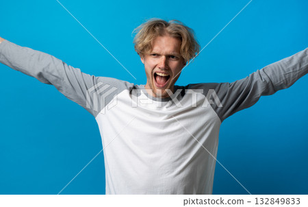 Triumphant man in gray and white shirt raising arms and shouting joyfully on blue background Triumphant man in gray and white shirt raising arms and shouting joyfully on blue background 132849833