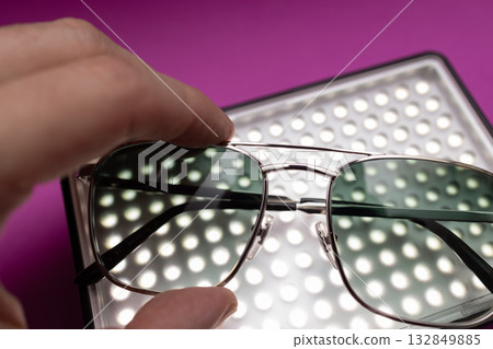 Hand holding silver frame sunglasses with smoky green lenses above LED panel on purple backdrop 132849885