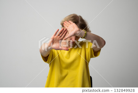 Man in yellow shirt covering face with hands showing fear and avoidance during photo session Man in yellow shirt covering face with hands showing fear and avoidance during photo session 132849928
