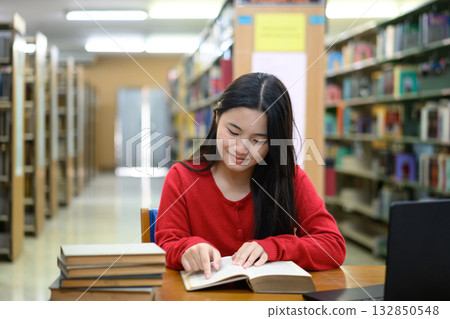 Asian teenage girl reading book with focus and smile in library study area 132850548