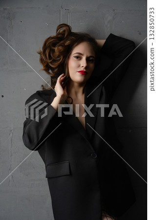 Confident woman posing elegantly in black blazer against a gray wall in a stylish indoor setting 132850733