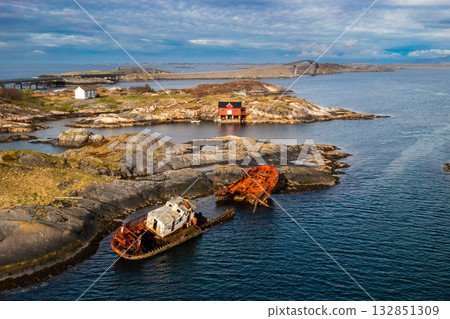 Rusty shipwreck submerged in waters off contrasts with red boathouse on islet. Atlantic ocean road hints at vast and rugged landscape in Norway 132851309