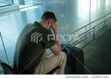 Young man with suitcase sitting in airport terminal looking upset, stressed and disappointed. Concept of flight delay, cancellation or travel problems. 132851342