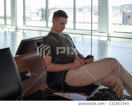 Young man charging phone at airport gate, using smartphone while waiting for flight. Comfortable travel concept in modern terminal. 132851343
