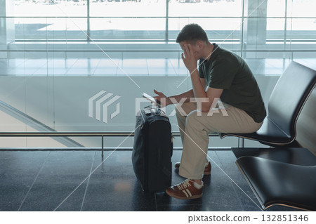 Young man sitting at airport gate with suitcase waiting for a flight, looking at his smartphone. Passenger is upset with flight delay. Modern air travel concept. 132851346