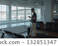 Man stands at a large airport window, holding his suitcase and checking his phone while waiting for his flight. Concept of solitude, anticipation and modern travel technology. 132851347