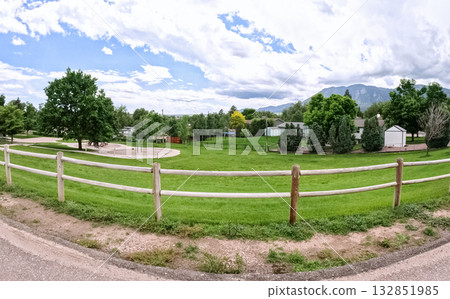 A wide open field surrounded by a white wooden fence, with mature trees and distant mountains under a bright sky. The image captures a peaceful, rural edge of a suburban neighborhood. 132851985