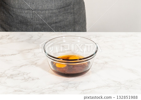 Side view of a glass bowl with eggs and maple syrup, placed on a marble countertop as part of the Amber Gingerbread Cookie Dough preparation, highlighting the combination of wet ingredients for the Side view of a glass bowl with eggs and maple syrup, placed on a marble countertop as part of the Amber Gingerbread Cookie Dough preparation, highlighting the combination of wet ingredients for the 132851988