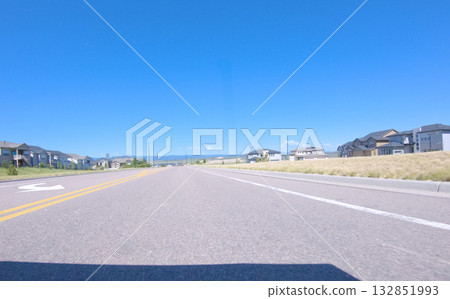 Eye-level view of a wide road in a residential area lined with modern houses. The road stretches ahead under clear blue skies, creating a clean and open suburban landscape. 132851993