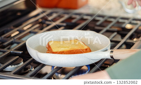 Within the sleek setting of a modern white kitchen, banana slices and brioche bread sizzle on a non-stick pan over a gas stove, setting the stage for an enticing grilled peanut butter banana sandwich. 132852002