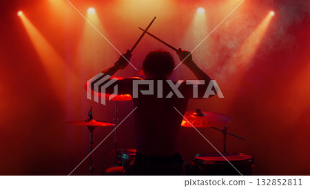 Drummer silhouette under orange light raising drumsticks in emotional finale 132852811