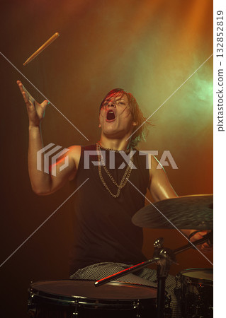 Drummer tossing drumstick in air with expression and warm stage lighting Drummer tossing drumstick in air with expression and warm stage lighting 132852819
