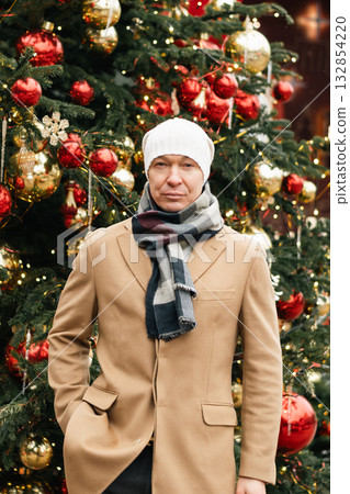 Man in cozy winter outfit stands outdoors near decorated Christmas tree, showcasing holiday decorations. Festive spirit of Xmas holiday with light garland. Man in cozy winter outfit stands outdoors near decorated Christmas tree, showcasing holiday decorations. Festive spirit of Xmas holiday with light garland. 132854220