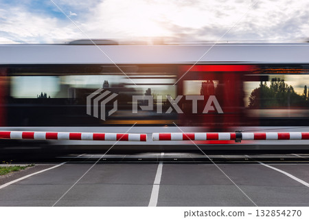Fast modern regional commute train rushes past closed railway crossing under cloudy sky sunlight sunset evening. Motion blur rush hour speed dynamic atmosphere urban transportation commute scene Fast modern regional commute train rushes past closed railway crossing under cloudy sky sunlight sunset evening. Motion blur rush hour speed dynamic atmosphere urban transportation commute scene 132854270