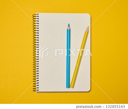 A spiral-bound notebook with blank pages, topped with blue and yellow pencils, sits against a yellow background. 132855143