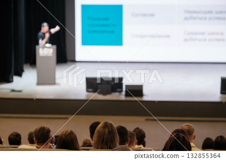 Energetic speaker presents at conference to attentive audience in a large hall with a bright screen Energetic speaker presents at conference to attentive audience in a large hall with a bright screen 132855364