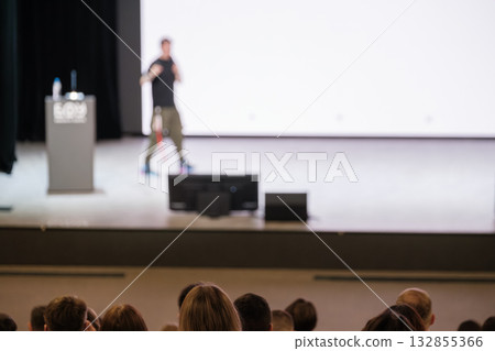 Speaker on stage delivering presentation to audience with podium and large screen 132855366