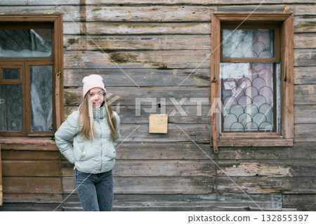 Young woman in mint jacket posing near old wooden house. Contrast between youth, texture, and rustic architectural background with copy space 132855397