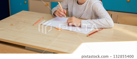 Child drawing in notebook with orange pencil during classroom lesson, banner with copy space. Creativity, imagination, and focus on self-expression through learning. 132855400