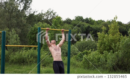 Muscular athlete doing pull ups on horizontal bar at sports ground. Young shirtless sportsman exercising at nature. Strong sporty man training outdoor. Concept of healthy and active lifestyle. Slow mo Muscular athlete doing pull ups on horizontal bar at sports ground. Young shirtless sportsman exercising at nature. Strong sporty man training outdoor. Concept of healthy and active lifestyle. Slow mo 132855465