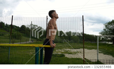 Young sportsman doing push ups on parallel bars at nature. Strong sporty man exercising at sports ground. Male shirtless athlete training hard outdoor. Concept of active sportive lifestyle. Slow mo Young sportsman doing push ups on parallel bars at nature. Strong sporty man exercising at sports ground. Male shirtless athlete training hard outdoor. Concept of active sportive lifestyle. Slow mo 132855466