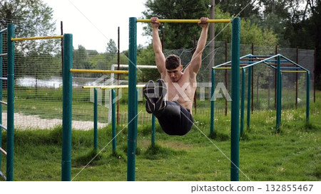 Athletic man trains abdominal muscles raising legs on horizontal bar at sports ground. Young sportsman exercising on pull-up bar outdoor. Sporty athlete working out at nature. Healthy lifestyle 132855467