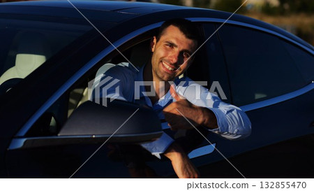 Young smiling entrepreneur leaning out of driver window at modern electric car and looking into camera. Portrait of handsome successful businessman showing thumbs up after bought new electrical auto Young smiling entrepreneur leaning out of driver window at modern electric car and looking into camera. Portrait of handsome successful businessman showing thumbs up after bought new electrical auto 132855470