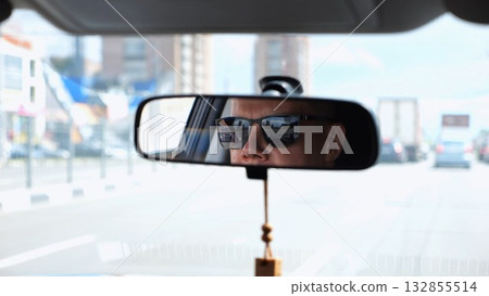Eyes in sunglasses of male driver reflecting in rearview mirror while riding motor auto. Young guy looking concentrated to urban road during operating vehicle. Confident man driving modern car at city Eyes in sunglasses of male driver reflecting in rearview mirror while riding motor auto. Young guy looking concentrated to urban road during operating vehicle. Confident man driving modern car at city 132855514