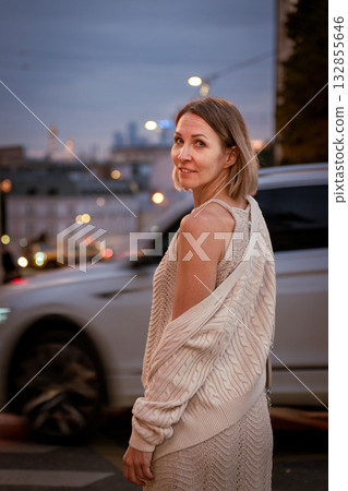 Woman in a beige knitted outfit stands on a city street at twilight, turning back with a gentle smile 132855646